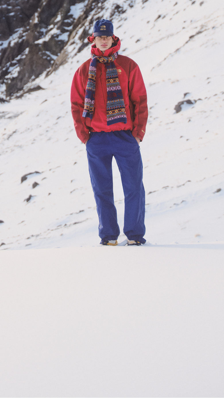 Man in red jacket, jacquard scarf and blue pants against a snowy mountain backdrop.