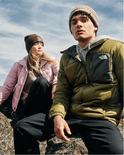 Two people are sitting outside, one is wearing a pink jacket and the other one is wearing a green jacket.