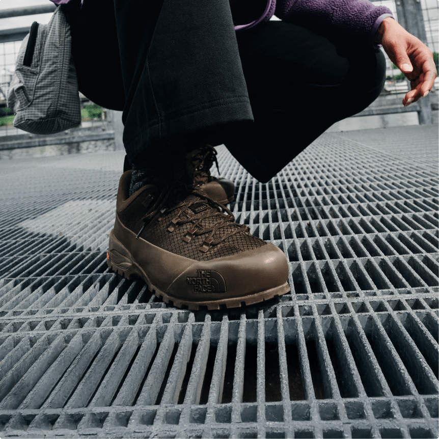 Gros plan d’une chaussure brune et de la jambe d’une personne portant un pantalon noir, accroupie sur une surface métallique grillagée.