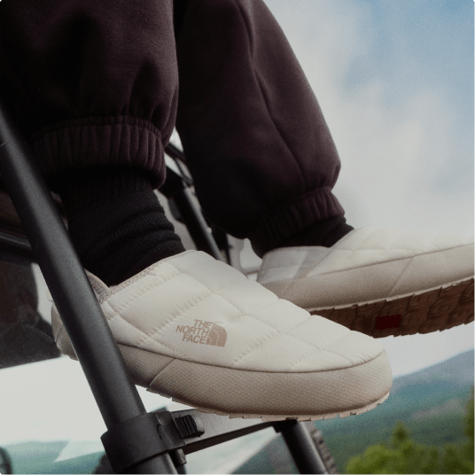 Closeup of a person’s white, padded slip-on shoes hanging in the air, mountains in the background. 