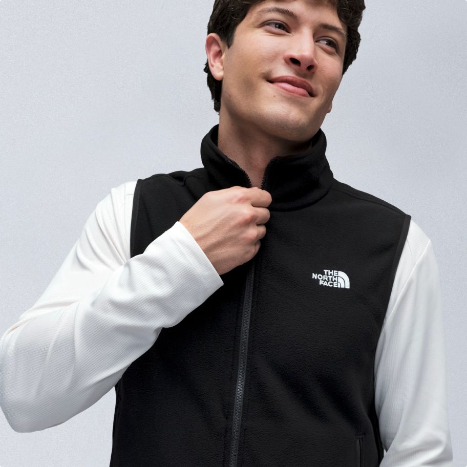 Man with brown hair wearing black fleece vest over a white longsleeve t-shirt, with his hand at the top of the zipper.