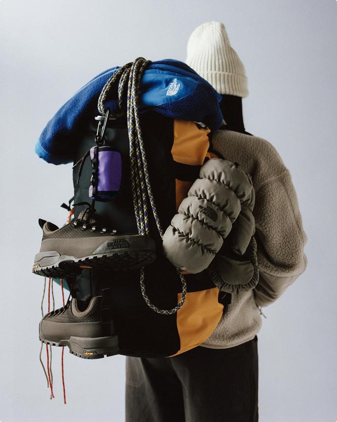 Back of person wearing a white beanie, holding a pile of gear starting with a blue jacket, gray pack and gray shoes slung over their pack with a white knit beanie on.