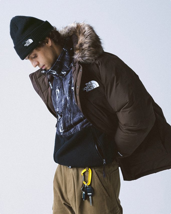 Man wearing black cuffed beanie, brown fur-trim hood jacket that’s open, printed mock-neck with black pouch pocket, yellow carabiner and khaki pants.