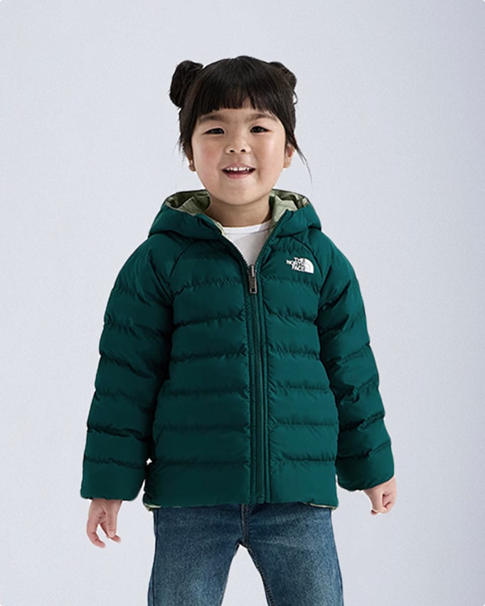 Kid with black bangs and hair in pigtail buns, green hooded zip-up jacket, white t-shirt poking through and jeans.