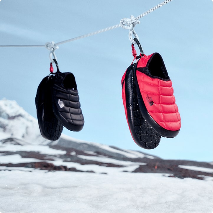 Sky-blue background with white mountain tops on the left, white rope hanging with Thermoball mules in black, hot pink and purple on carabiners.