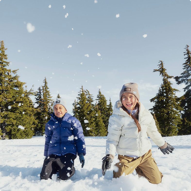 Tree-lined background with clear blue sky, boy on left wearing blue jacket and black pants, girl on right wearing white jacket and beige pants, both kneeling on the snow having a snowball fight.