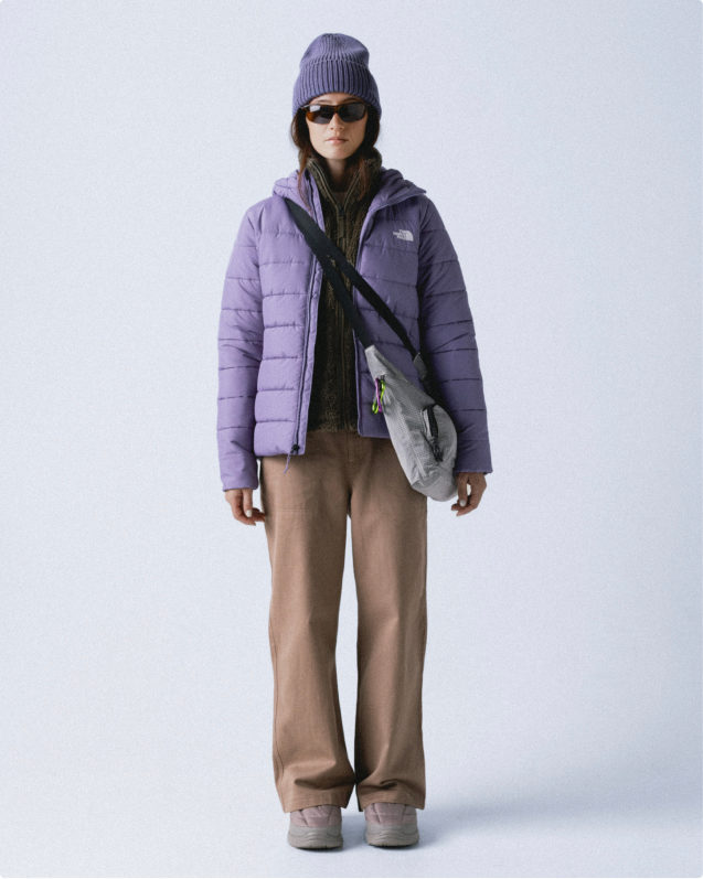 Woman wearing purple knit beanie, unzipped hooded purple puffer jacket, light gray crossbody bag and khaki pants.