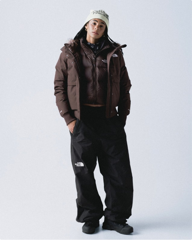 Woman wearing white beanie, unzipped dark plum fur-trim hooded jacket, zipped-up brown puffer underneath and black pants.