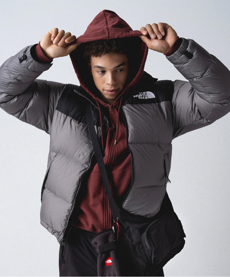 Man wearing maroon hooded zip-up with hood on, with gun-metal gray/black retro Nuptse jacket unzipped and a black crossbody bag.