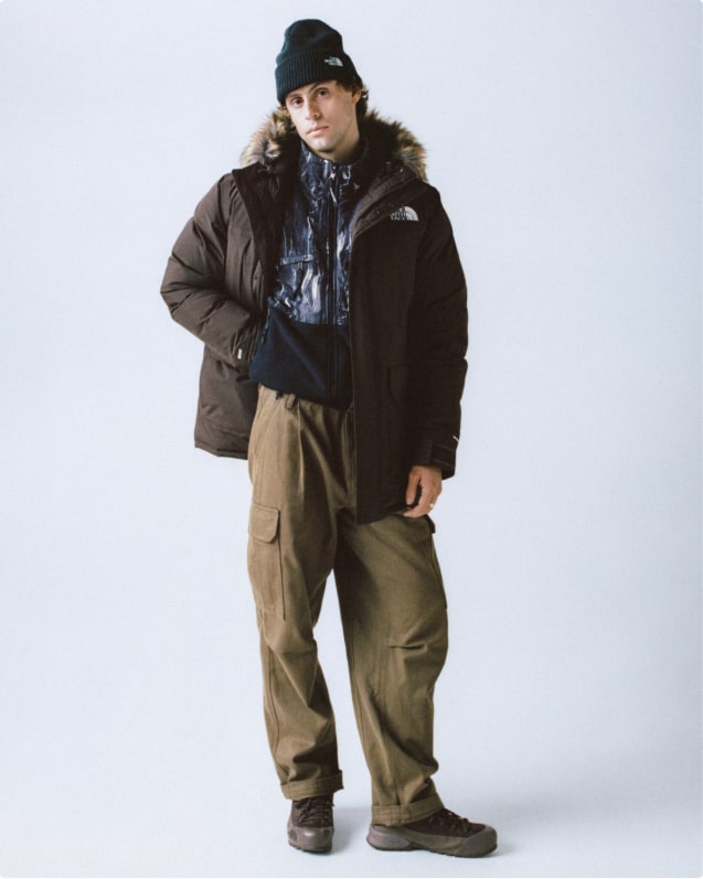 Man wearing black cuffed beanie, brown fur-trim hood jacket that’s open, printed mock-neck with black pouch pocket, yellow carabiner and khaki pants.