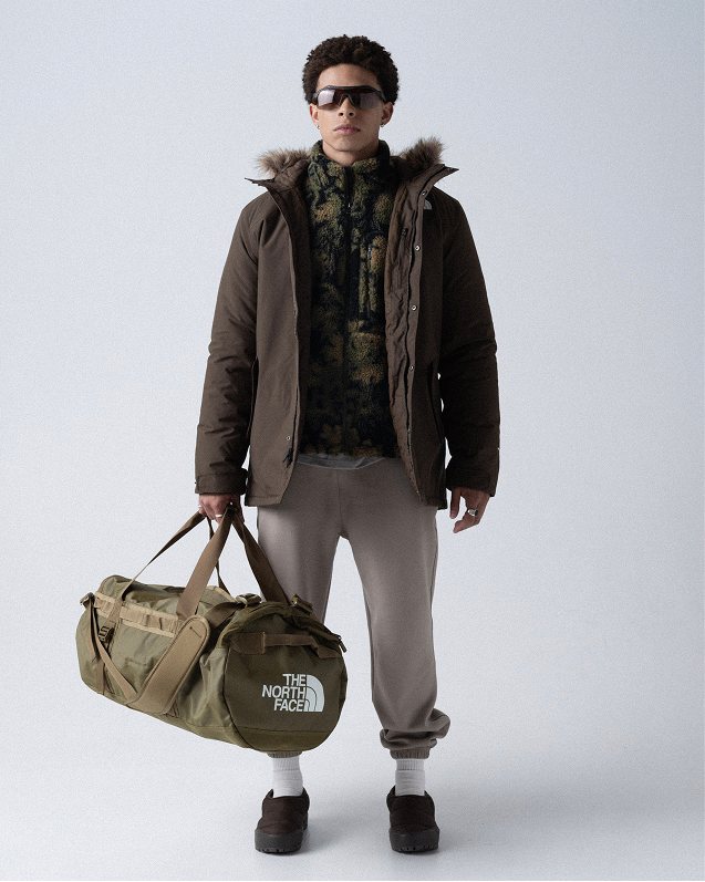 Man wearing black sunglasses, unzipped fur-trim hooded jacket, camo print fleece underneath, gray sweatpants, white socks and black shoes, holding an army-green duffel bag.