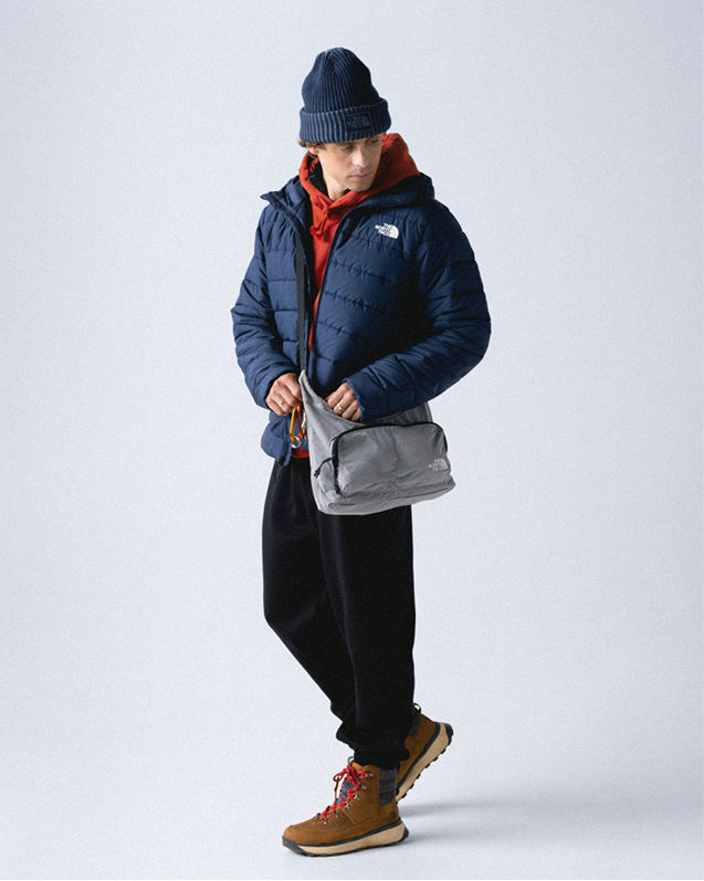Man wearing a navy cuffed beanie, red hoodie, navy unzipped puffer jacket, gray crossbody bag, black sweatpants and brown boots with red laces.
