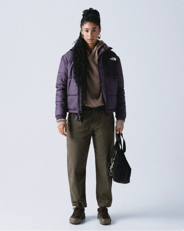 ImageWoman with purple jacket unzipped, oatmeal thermal underneath, and dark gray pants, holding a black bag.