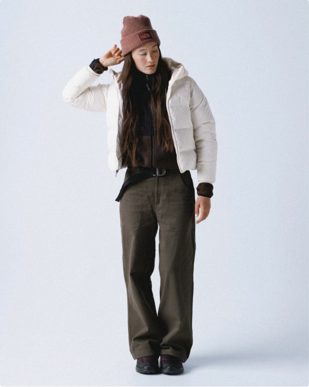 Woman wearing mauve cuffed beanie, white open puffer jacket, brown zipped-up fleece beneath and dark gray wide-leg pants.
