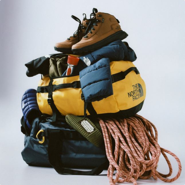 Pile of gear starting from top: brown boots, teal puffer, yellow duffel, pink rope on the right, all on top of a teal bag with a gold caribiner