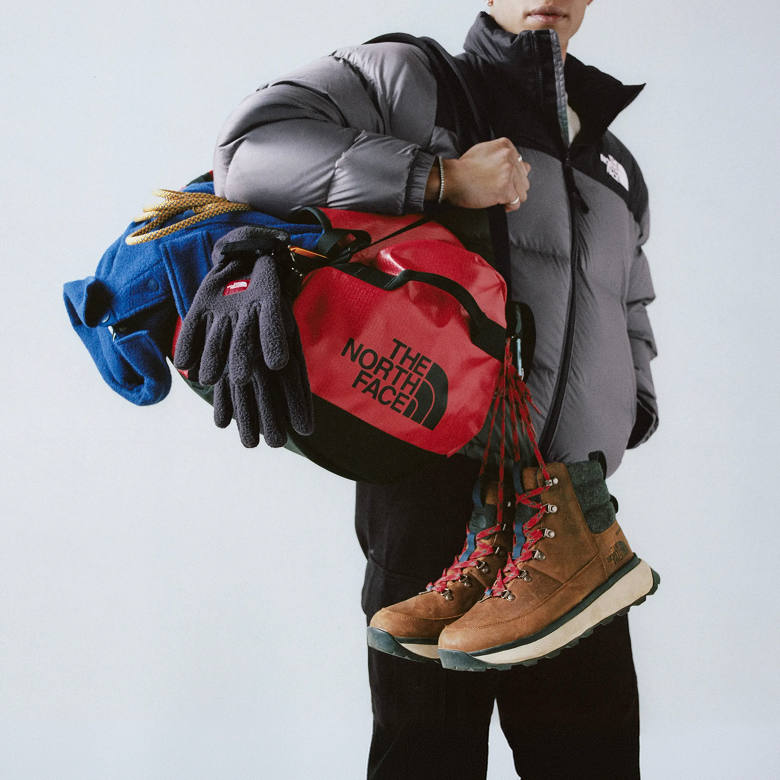 Man wearing the grey/black puffer jacket, holding a red duffel bag on his right arm with gear piled on: blue jacket, dark grey gloves and brown boots with red laces.