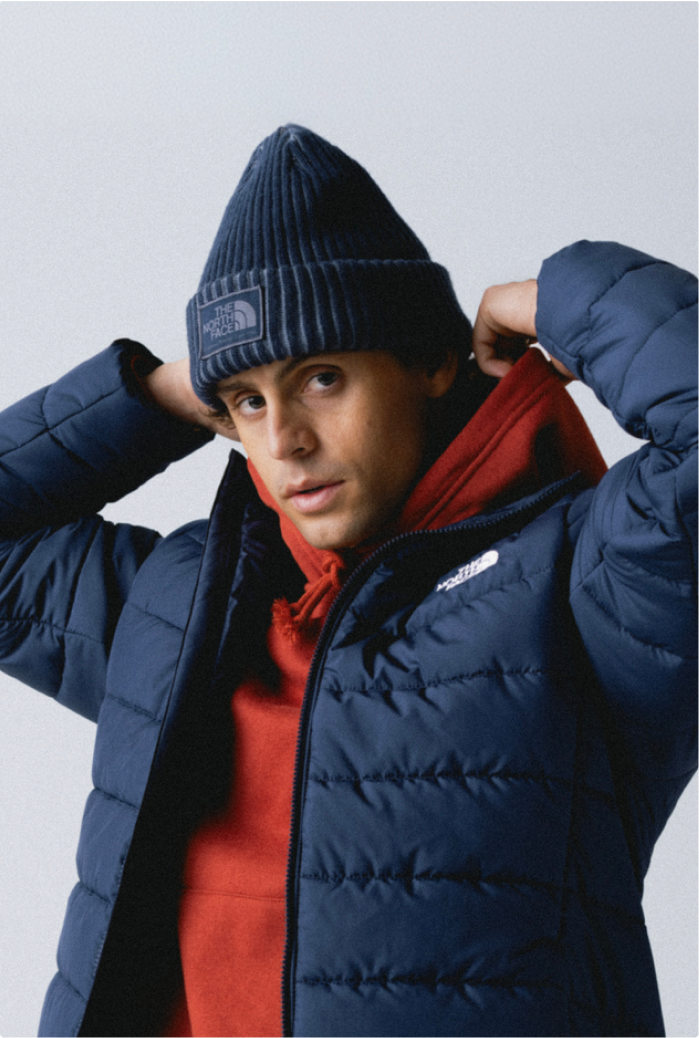 Man wearing blue cuffed beanie, red hoodie underneath an unzipped blue jacket. His arms are up holding the hood of his sweatshirt.