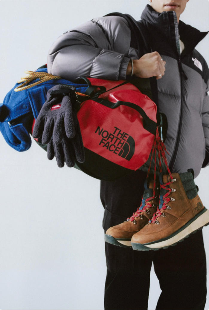 Man wearing the grey/black puffer jacket, holding a red duffel bag on his right arm with gear piled on: blue jacket, dark grey gloves and brown boots with red laces.