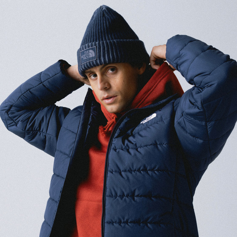 Man wearing blue cuffed beanie, red hoodie underneath an unzipped blue jacket. His arms are up holding the hood of his sweatshirt.