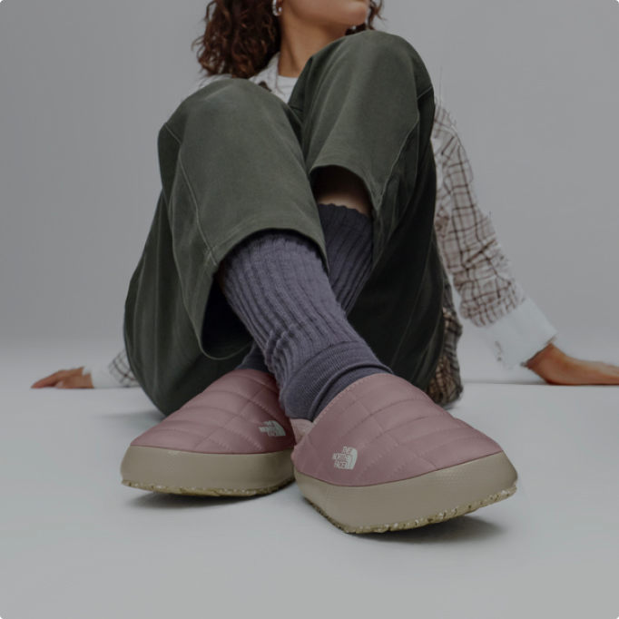 A woman sitting on the ground with her ankles crossed wearing pink Thermoball mules