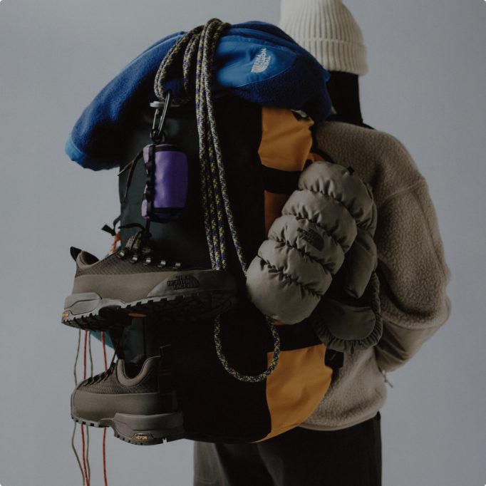 Back of person wearing a white beanie, holding a pile of gear starting with a blue jacket, gray pack and gray shoes slung over their pack with a white knit beanie on.