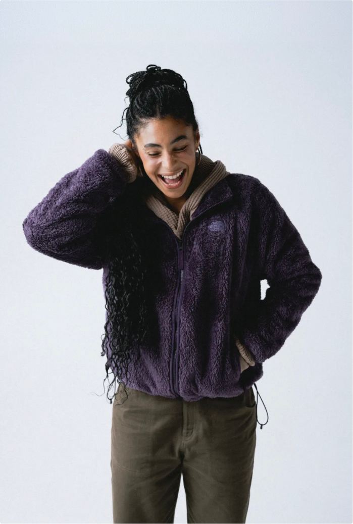 Woman smiling wide with her hair up, wearing a zipped-up purple fleece jacket with oatmeal thermal underneath and dark green/grey pants.