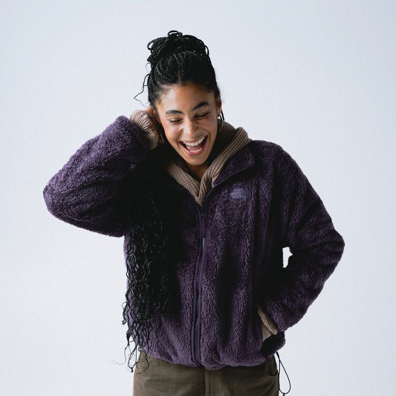 Woman smiling wide with her hair up, wearing a zipped-up purple fleece jacket with oatmeal thermal underneath and dark green/grey pants.