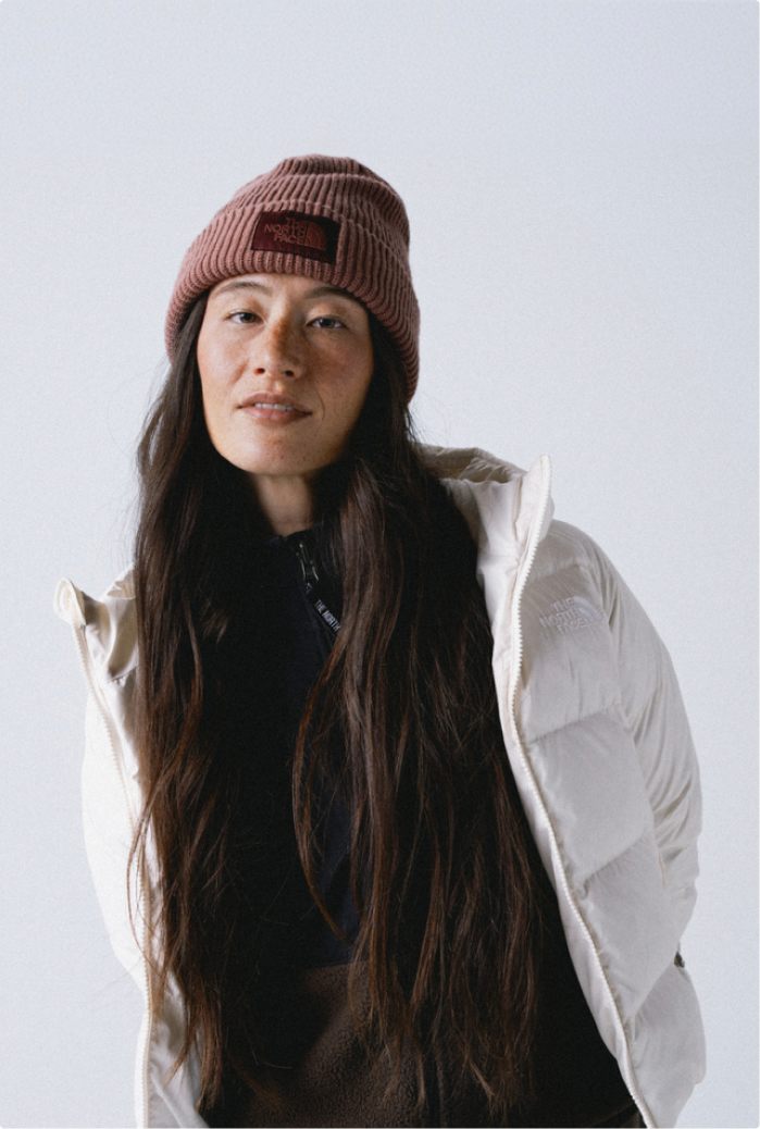 Woman wearing mauve cuffed beanie with her long brown hair down in front of her, wearing a black mock neck and an open white hooded jacket.