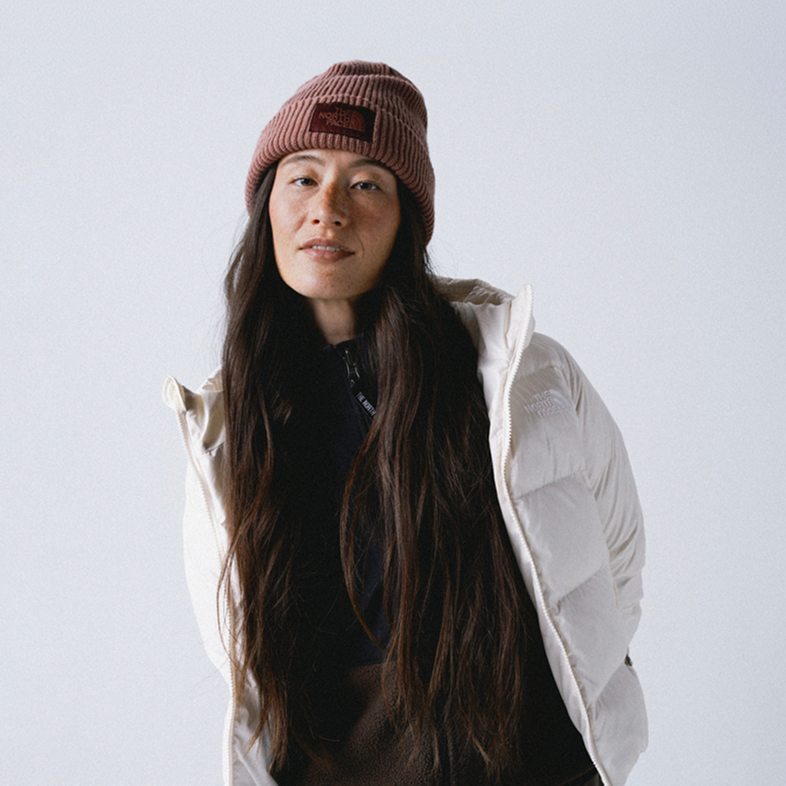 Woman wearing mauve cuffed beanie with her long brown hair down in front of her, wearing a black mock neck and an open white hooded jacket.