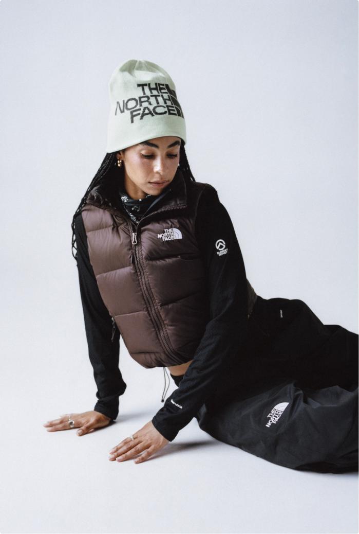Woman leaning on her side with her hands down, wearing an off white TNF beanie with a brown puffer vest, black long sleeve underneath, and black pants.