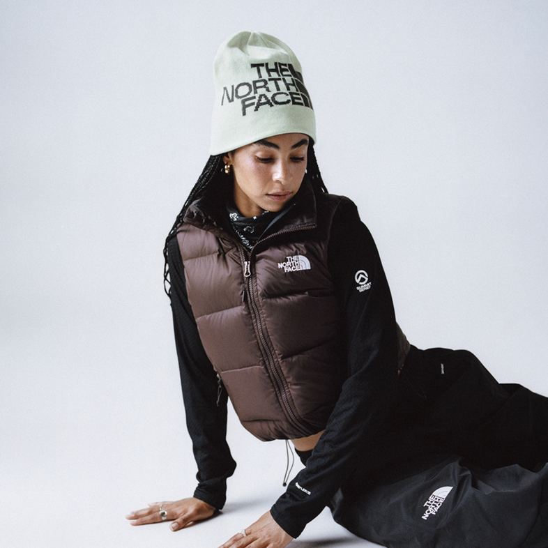 Woman leaning on her side with her hands down, wearing an off white TNF beanie with a brown puffer vest, black long sleeve underneath, and black pants.