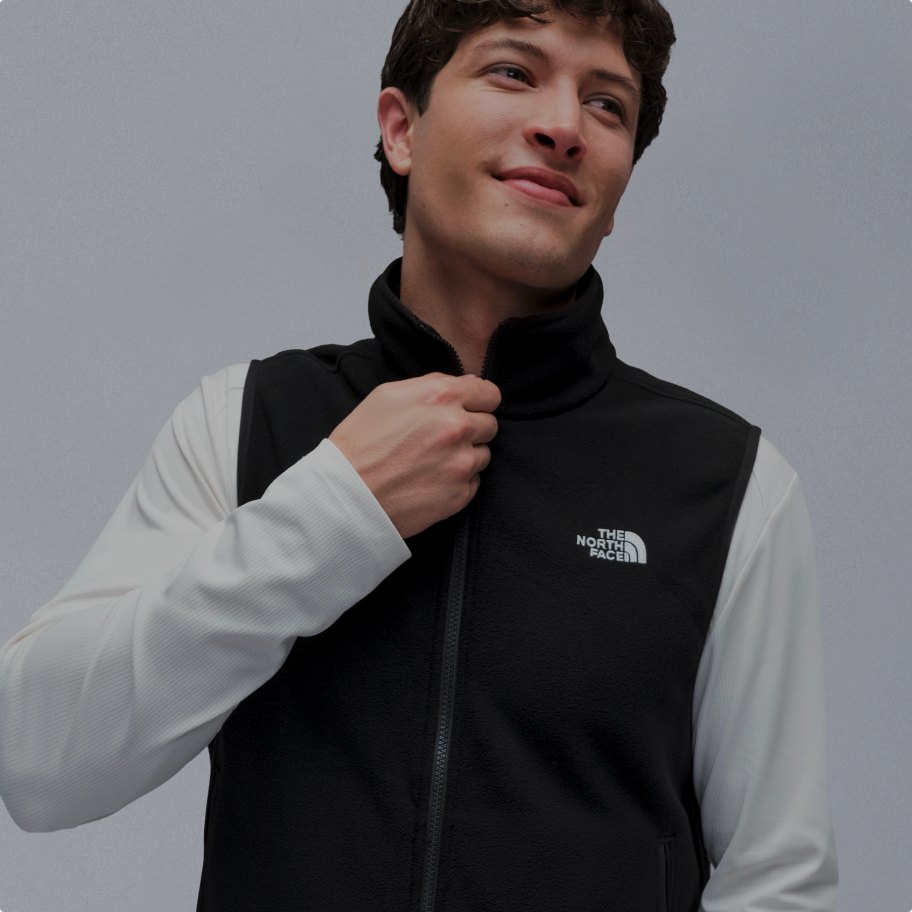 Man with brown hair wearing black fleece vest over a white longsleeve t-shirt, with his hand at the top of the zipper.