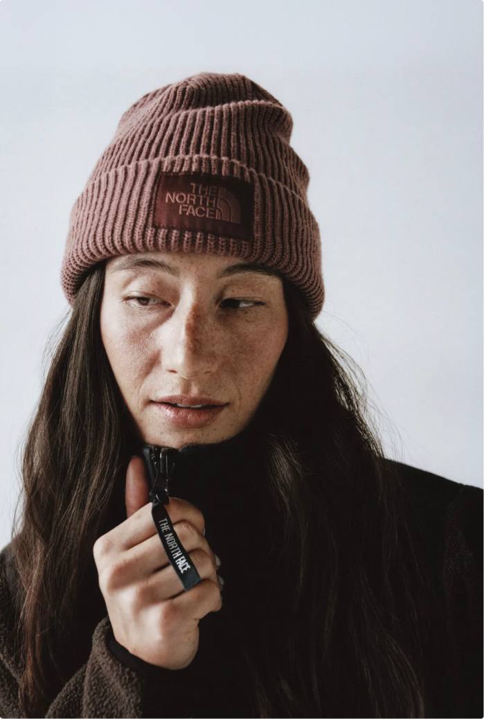 Close-up of woman wearing mauve cuffed beanie with her brown hair around her shoulders. She’d wearing a mock neck zip-up and her right hand is on the top of the collar.