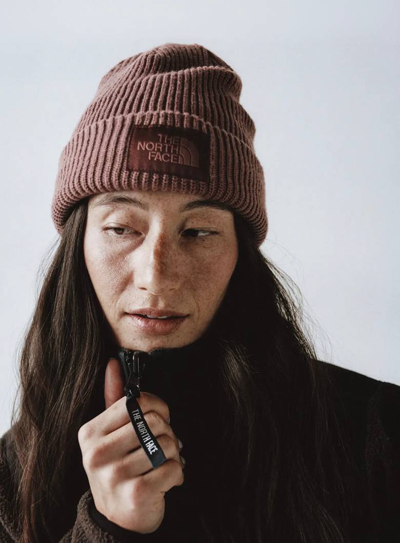 Close-up of woman wearing mauve cuffed beanie with her brown hair around her shoulders. She’d wearing a mock neck zip-up and her right hand is on the top of the collar.