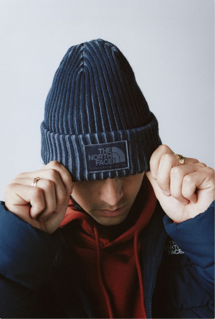 Man wearing a blue cuffed beanie down over his eyes, with a red hoodie and open blue jacket, with his fingers up on the edge of the cuffed beanie.