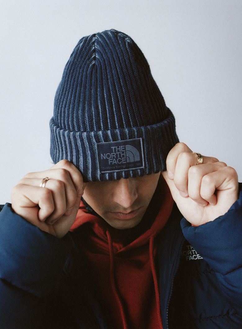 Man wearing a blue cuffed beanie down over his eyes, with a red hoodie and open blue jacket, with his fingers up on the edge of the cuffed beanie.