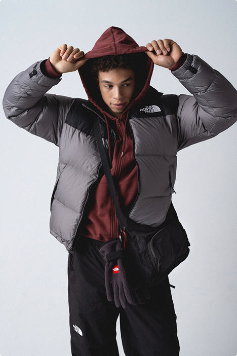 Man wearing maroon hooded zip-up with his hands on the hood, with gun-metal gray/black retro Nuptse jacket unzipped, a black crossbody bag and mountain gloves hanging from his waist.