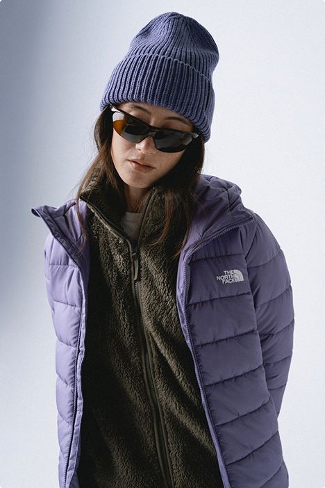 Woman wearing a purple beanie, black sunglasses, dark green fleece zipped up and purple puffer unzipped and open on her.
