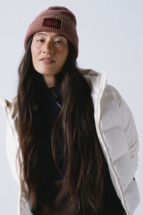 Woman with mauve beanie and long brown hair wearing a zipped-up black fleece and an open unzipped white jacket against white background.