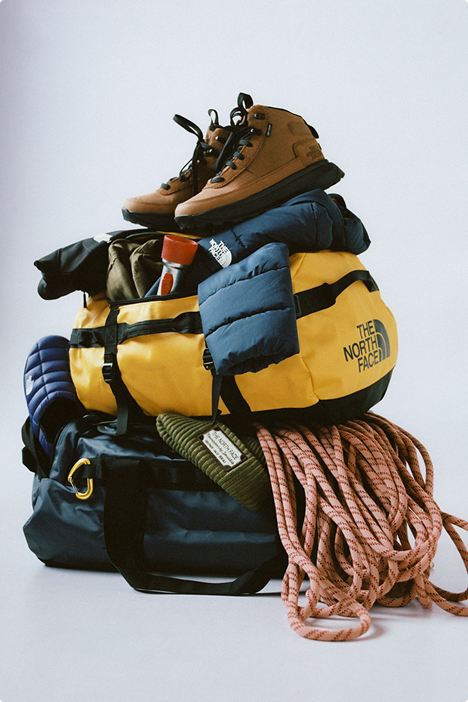 Pile of gear against a white background, with brown boots on top, then blue puffer, yellow duffel, pink rope all on top of a teal bag with a gold caribiner on the left.