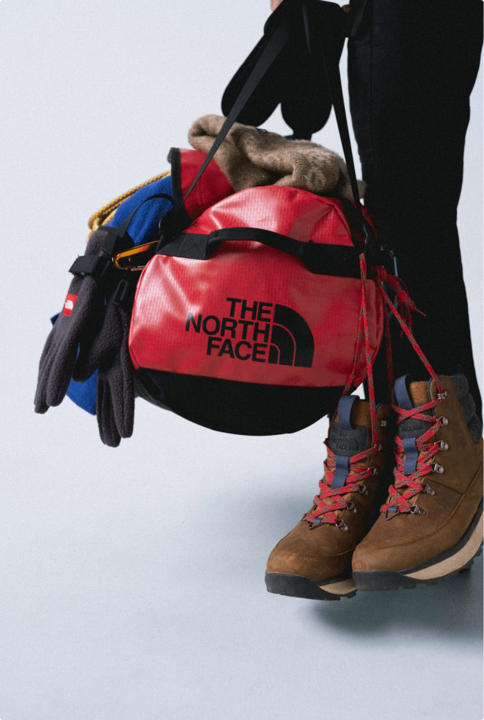 Person in black pants holding a red duffel bag with a pile of gear hanging off it: brown boots with red laces down the right, black gloves and blue jacket down the left.