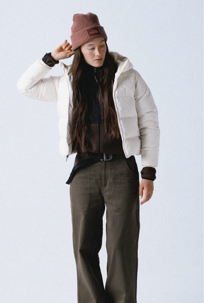 Woman wearing a mauve cuffed beanie and white insulated hooded jacket unzipped over a dark brown fleece and she’s wearing dark olive pants. Her arm is up and she’s holding the edge of the left side of the beanie.
