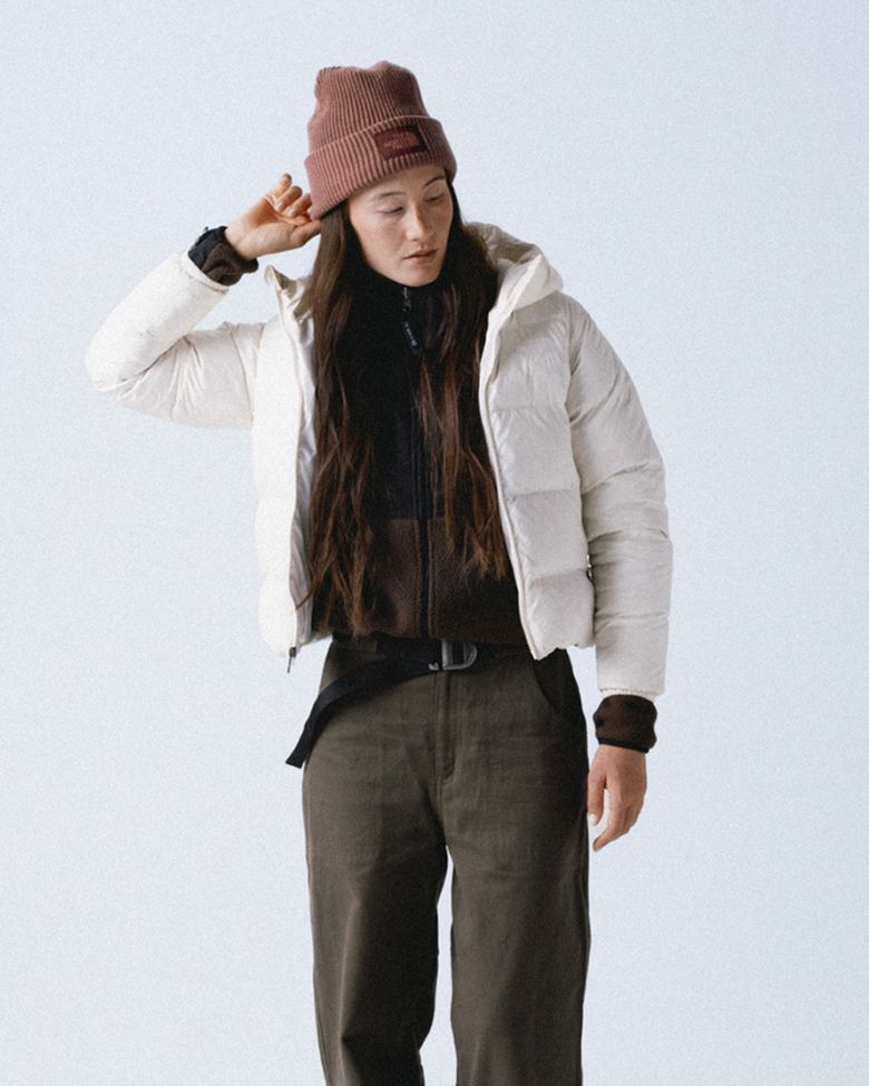 Woman wearing a mauve cuffed beanie and white insulated hooded jacket unzipped over a dark brown fleece and she’s wearing dark olive pants. Her arm is up and she’s holding the edge of the left side of the beanie.