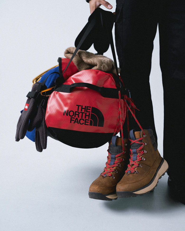 Person in black pants holding a red duffel bag with a pile of gear hanging off it: brown boots with red laces down the right, black gloves and blue jacket down the left.