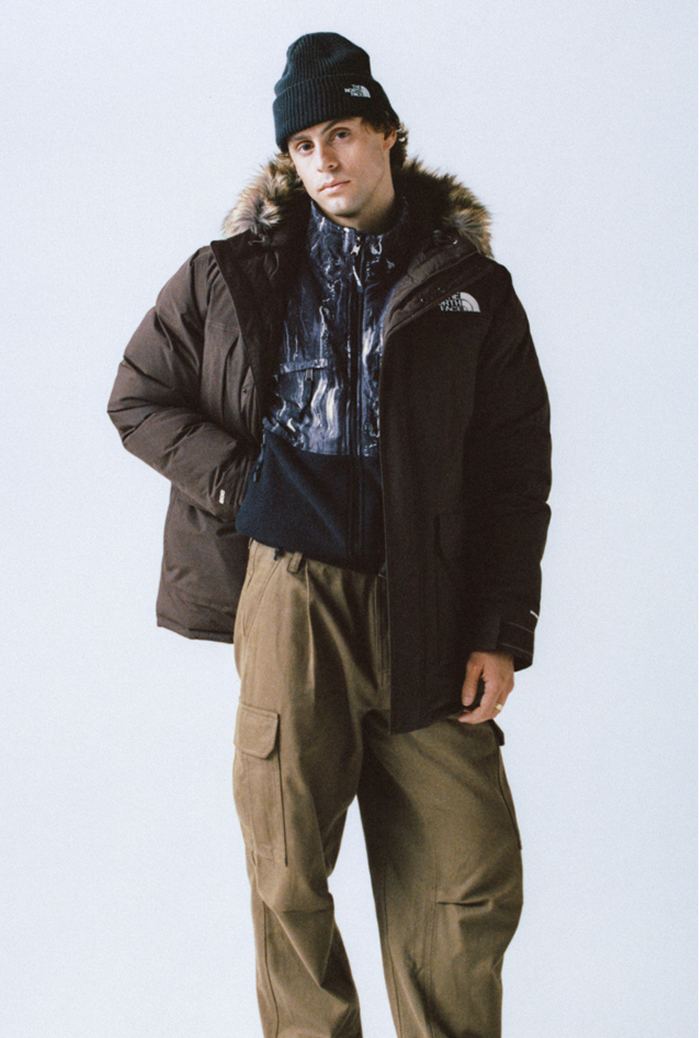 Man wearing a black cuffed beanie, the fur-trimmed brown jacket unzipped and open, with a printed mock neck underneath and khaki pants.
