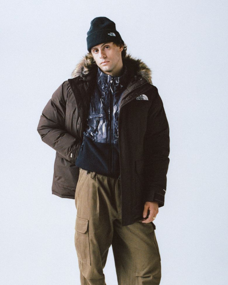 Man wearing a black cuffed beanie, the fur-trimmed brown jacket unzipped and open, with a printed mock neck underneath and khaki pants.