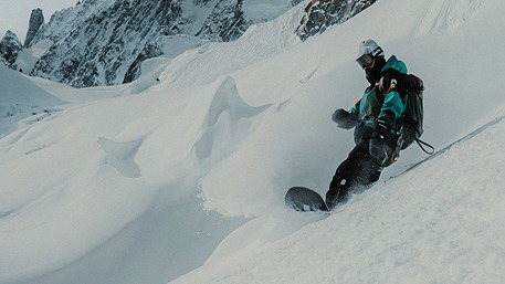 A woman in a black helmet and yellow reflective goggles skiing down powdery moguls. Her poles are gently sticking out on either side.