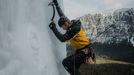 An ice climber in a yellow and black jacket climbing a rock face while sinking their left pick into a sheet of ice. They have a white helmet on.