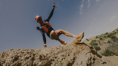 A woman in a white head band, turquoise jacket and black pants running up a rocky scramble.