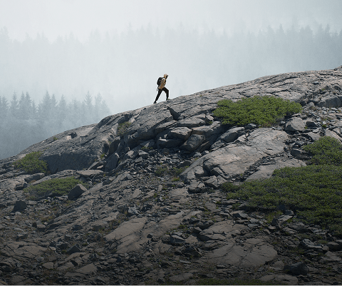 A person in a rain jacket from The North Face hikes up a rocky ridge. In the background, gray mist covers the trees.